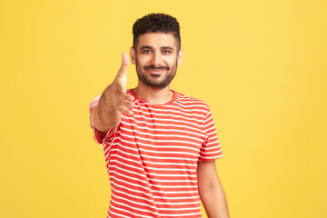 Fototapeta premium Nice to meet you. Positive friendly man with beard in striped t-shirt giving hand for handshaking, welcoming partners, job offer. Indoor studio shot isolated on yellow background