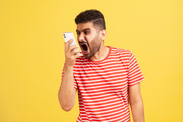 Angry mad man with beard in striped t-shirt loudly screaming talking phone, leaving voice message with furious aggressive expression. Indoor studio shot isolated on yellow background