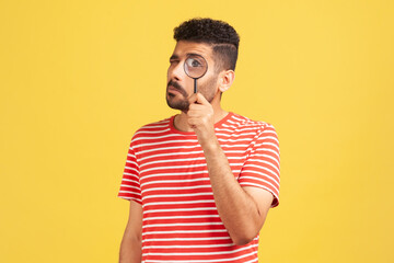 Serious bearded man in red striped t-shirt standing, holding magnifying glass and looking at camera...