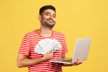Complacent self confident man smiling holding in hands laptop and fan of hundred bills, proud of...