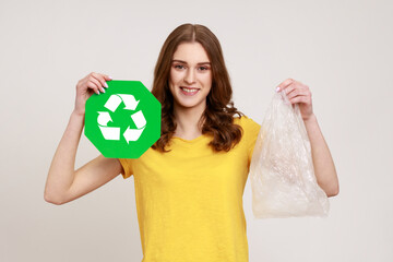 Portrait of beautiful happy teenager girl with brown hair wearing yellow casual T-shirt, holding in hands plastic package and green recycle sign. Indoor studio shot isolated on gray background.