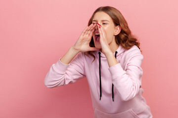 Portrait of annoyed aggressive teen girl holding hands shaped as megaphone and screaming frantically, proclaiming loud message. Indoor studio shot isolated on pink background