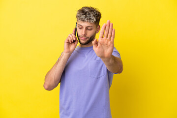 Naklejka premium Young caucasian man using mobile phone isolated on yellow background making stop gesture and disappointed