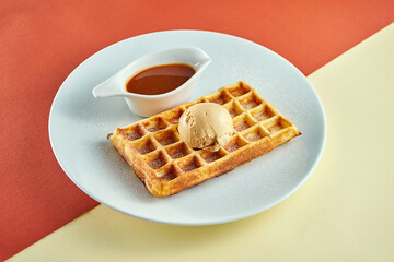 Classic Belgian waffles with ice cream and sweet sauce in a white plate on colored backgrounds.