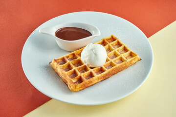 Classic Belgian waffles with ice cream and sweet sauce in a white plate on colored backgrounds.
