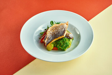 Fried sea bass fillet with steamed vegetables for garnish in a white plate on colored backgrounds. - obrazy, fototapety, plakaty