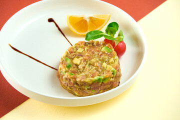 Salmon tartare with avocado and lemon in a white plate on colored backgrounds.