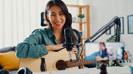 Teenage Asia girl influencer play guitar music use microphone record with smartphone for online audience listen at home. Female podcaster make audio podcast from her home studio, Stay at home concept.
