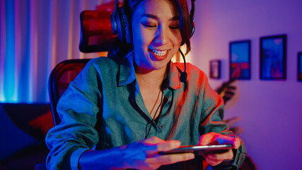 Happy asia girl gamer wear headphone competition play video game online with smartphone colorful neon lights in living room at home. Esport streaming game online, Home quarantine activity concept. © tirachard