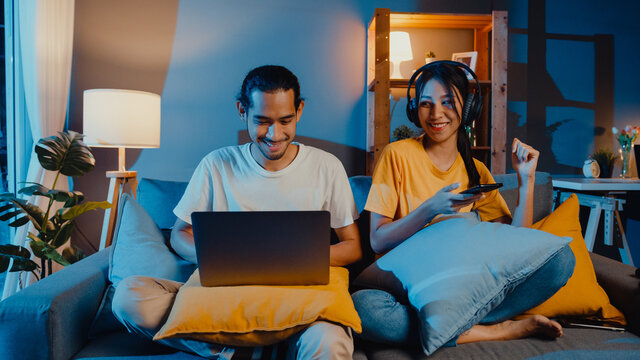 Freelance asia couple man and woman in casual hasband work  laptop computer at night enjoy happy relax with wife wear headphone listen music in smartphone sing dance on couch at house, Married life.