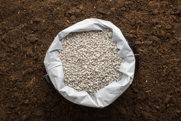 Opened plastic bag with gray complex fertiliser granules on dark soil background. Closeup. Product for root feeding of vegetables, flowers and plants. Top down view. - obrazy, fototapety, plakaty