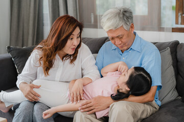 Asian grandparents talking with granddaughter at home. Senior Chinese, old generation, grandfather and grandmother using family time relax with young girl kid lying on sofa in living room concept.