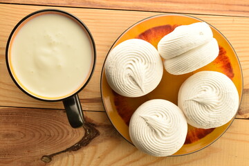 Several white sweet marshmallows on a ceramic saucer with a cup of milk, on a wooden table, close-up, top view.