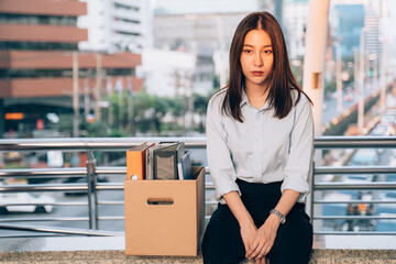 Stressed and worried young Asian woman with box of items sitting alone after being laid off from job due to recession and economic stress in industry - obrazy, fototapety, plakaty