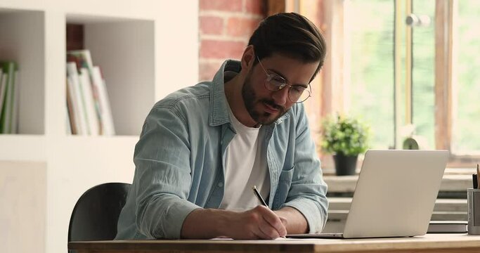 Concentrated young businessman in eyewear working on project online, typing in computer program, writing notes in paper document. Motivated millennial caucasian man studying distantly in office.