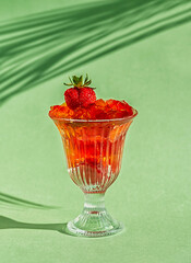 Ripe strawberries and delicious fruit jelly in a glass glass on a green background. Vertical crop. Close-up.