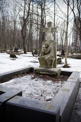 Stone cross on grave with statue of crying woman. In snowbound cemetery among leafless trees