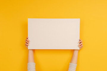 Hands holding white canvas paper blank with copyspace on yellow background. Empty sheet banner with...