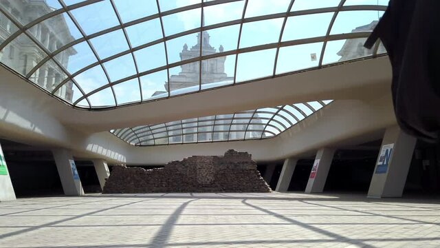 Woman tourist underground Serdica, glass dome and ancient historical ruins of the roman city, with view of the National Assembly building in the background. Slow motion
