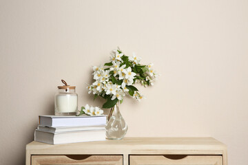 Bouquet of beautiful jasmine flowers in glass vase, books and candle on wooden commode near beige wall indoors - obrazy, fototapety, plakaty
