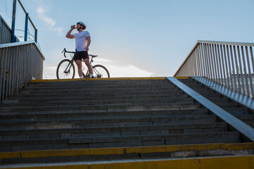 Low angle shot of a male muscular cyclist drinking water, standing near his bicycle on a summer day, copy space