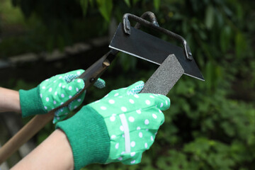 Woman sharpening hoe outdoors, closeup. Gardening tools - obrazy, fototapety, plakaty