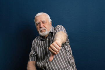 Charismatic gray-haired man showing thumb down, emotion in the frame, half-length portrait, blue background
