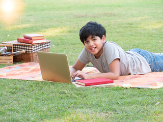Happy India boy plays with a laptop, lying on the grass at the park with a smiling face, kid, and technology concept..