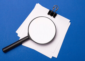 On a blue background, white papers under a black paper clip and on them a magnifying glass with...