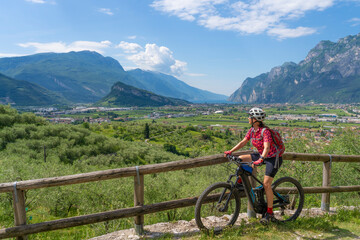 nice senior woman riding her  electric mountain bike in the olive groves of Arco and enjoying the awesome view over Garda Lake between Riva del Garda and Torbole - obrazy, fototapety, plakaty