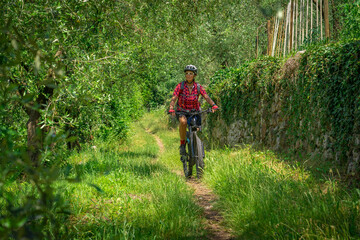 nice and active senior woman riding her electric mountain bike in the Garda lake mountains between blooming the olive groves of Arco close to Riva del Garda and Garda Lake , Landscape - obrazy, fototapety, plakaty