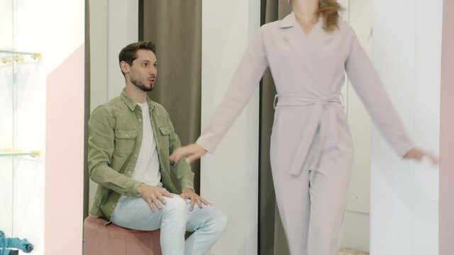 Young woman is trying on clothes in fitting room then showing garment to boyfriend smiling enjoying shopping together. Relationship and leisure toncept.