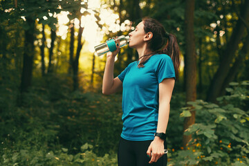 Photo of young athlete woman in sport wear drinking water while training in forest