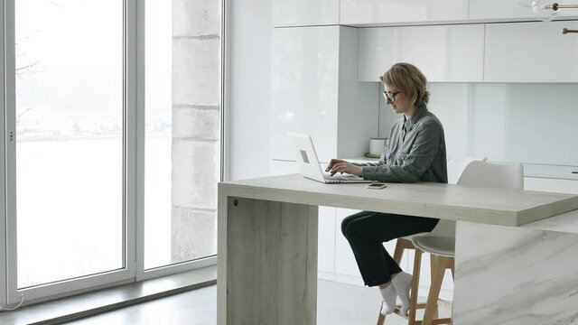 Professional businesswoman with short fair hair types on laptop sitting at wooden designed table in stylish room with french window