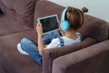 little cute girl with headphones and a tablet is sitting at home on the couch. The child is happy to learn at home online on a computer tablet, listening to music. A new children's learning app.