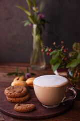 coffee with milk foam in a glass mug on a wooden table next to a bouquet of wildflowers and cookies