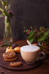 coffee with milk foam in a glass mug on a wooden table next to a bouquet of wildflowers and cookies