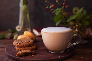 coffee with milk foam in a glass mug on a wooden table next to a bouquet of wildflowers and cookies