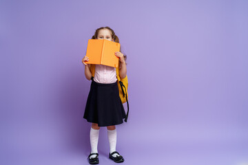 charming mischievous little girl in a school uniform with two ponytails smiles at the camera and hides behind a book, having fun during school classes on a purple background. Back to school.