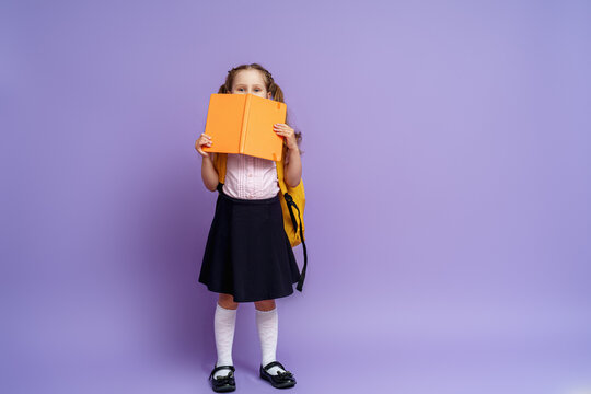 charming mischievous little girl in a school uniform with two ponytails smiles at the camera and hides behind a book, having fun during school classes on a purple background. Back to school.