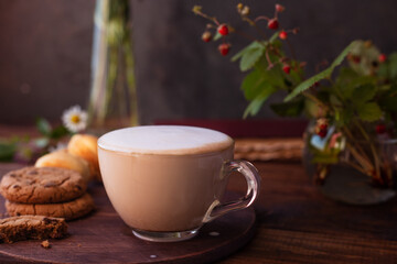 coffee with milk foam in a glass mug on a wooden table next to a bouquet of wildflowers and cookies