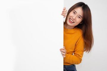 Happy young asian woman in yellow shirt standing behind blank white billboard