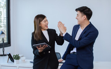 Two Asian handsome businessman and beautiful smart businesswoman wearing formal suits, making high five and tagging hands for unity, teamwork and successful projects, standing in indoor office.