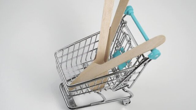 Wooden eco-friendly set of kitchen utensils with a spoon, fork and knife falls into a mini shopping cart on a white background in slow motion. Close-up. Ecology conservation concept