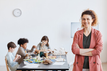 Waist up portrait of smiling female teacher looking at camera with group of children making wooden...