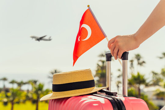 Woman with pink suitcase, hat and Turkey flag standing on passengers ladder and getting out of airplane opposite sea coastline with palm trees.