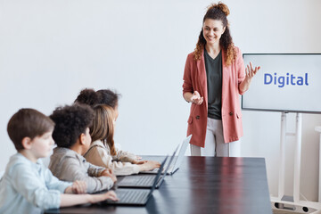 Portrait of smiling female teacher talking to group of kids using computers during IT lesson in school