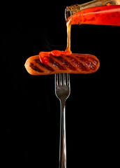 Fried sausage on a fork. Isolated on a black background. Cholesterol, junk food. Dynamics in the frame, pouring hot sauce ketchup on the grilled sausage. Selective Focus