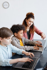 Vertical portrait of young female teacher helping boy using laptop during IT class in school