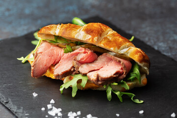 Fresh baked croissant sandwich with arugula and roast beef on a black slate board